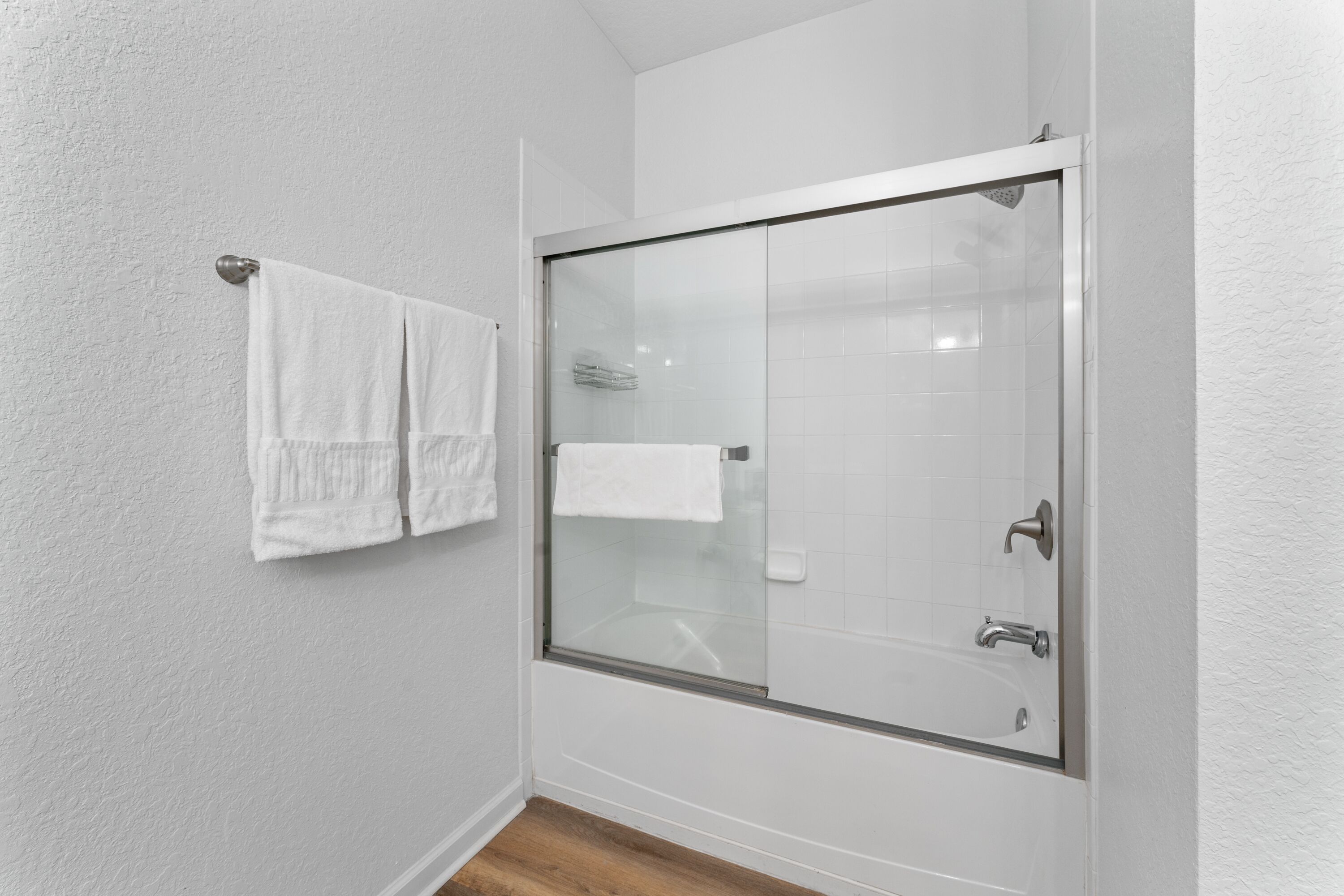 Une salle de bain dotée d’une douche à parois de verre, de murs recouverts de carreaux blancs et d’un plancher en bois.
