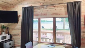 A cabin with a wooden interior, a dining table with chairs, a TV mounted on the wall, and a view of a lake through large windows. - Modern lakeside cabin with panoramic views and deck. (Alexandria)