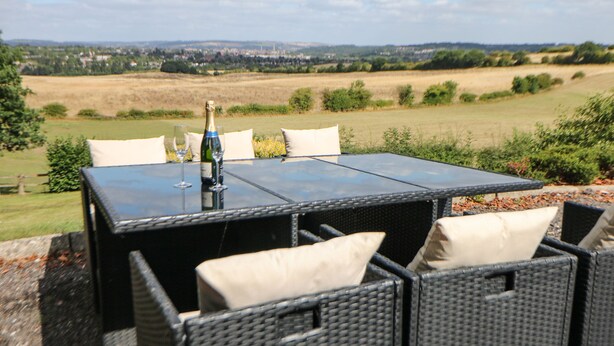 A table with a bottle and glasses, surrounded by wicker chairs with cushions, set against a view of a rural landscape. - Cedar Holme (Chesterfield)