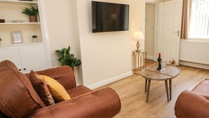 A living room with a brown sofa, a TV mounted on the wall, a small round table with a bottle and glasses, and a lamp on a side table. - Cedar Holme (Chesterfield)