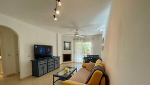 Ein Wohnzimmer mit einer Couch, einem Fernseher und einem Glastisch. Ein Kamin und ein Deckenventilator sind ebenfalls vorhanden.