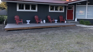 A house with a dark exterior, a red door, and a porch with four red Adirondack chairs and two white planters. - Cozy home next to airport with fenced backyard and lots of sun! (Juneau)