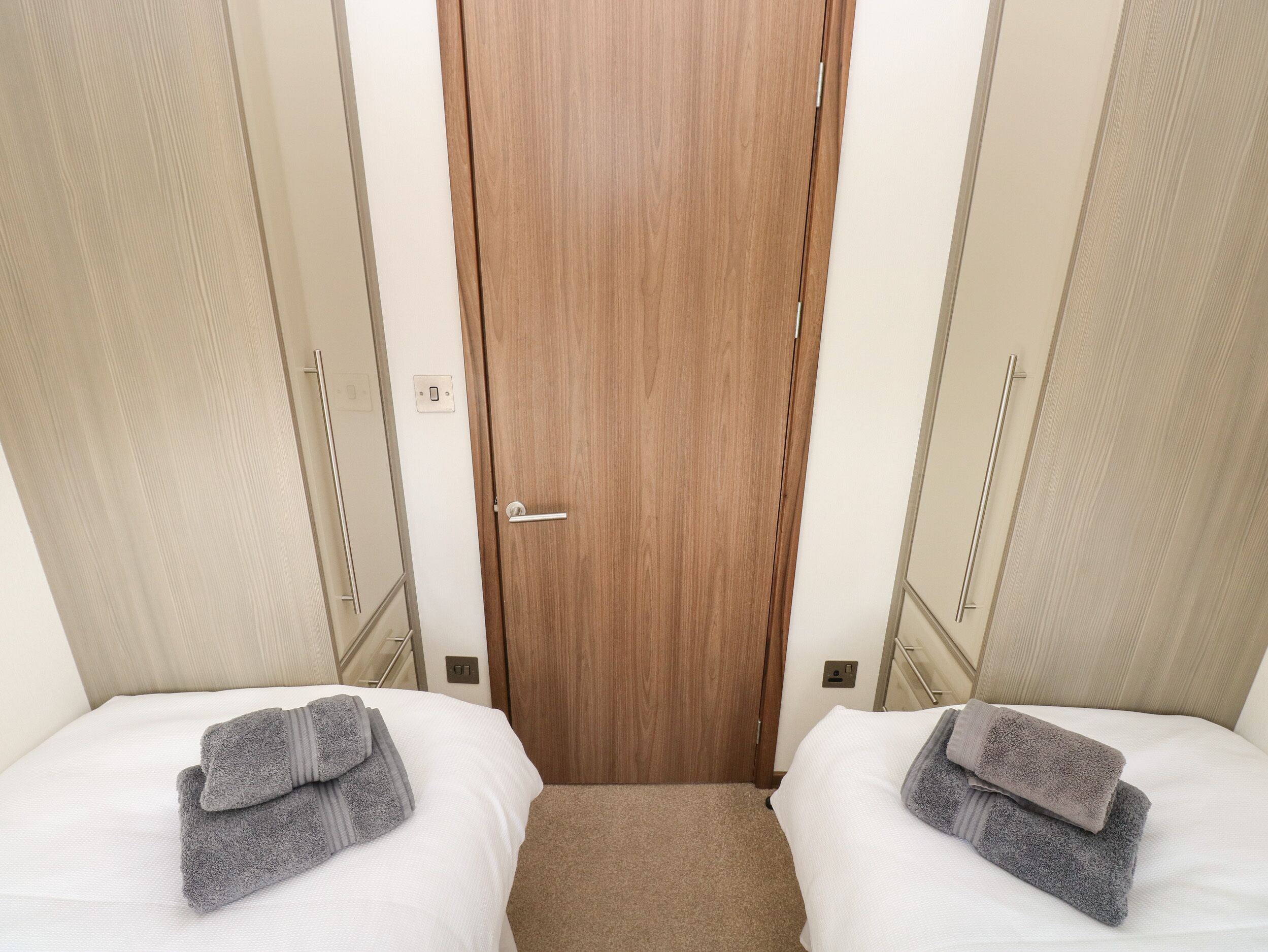 Une chambre d’hôtel avec deux lits, chacun agrémenté d’une serviette pliée, une porte en bois et un petit espace recouvert d’une moquette situé entre les lits.