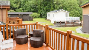 Une terrasse dotée de chaises en osier et d’une rambarde en bois, qui donne sur une zone verdoyante avec un bâtiment blanc et un quai.
