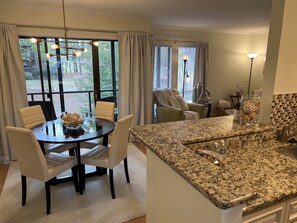 A modern kitchen with granite countertops, a dining area with a glass table, and a cozy seating area with a green armchair.