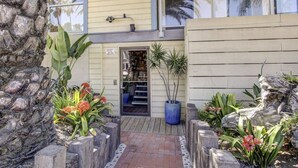 A two-story building with a beige exterior, numbered 2595, featuring a small entrance with a wooden door, a balcony, and a garden area with tropical plants and a large planter. - Ocean Front View - Steps from the beach! (San Diego)