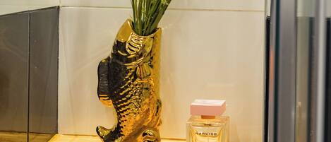 A bathroom with a golden fish-shaped vase containing yellow flowers and a bottle of Narsiso perfume on a wooden shelf.