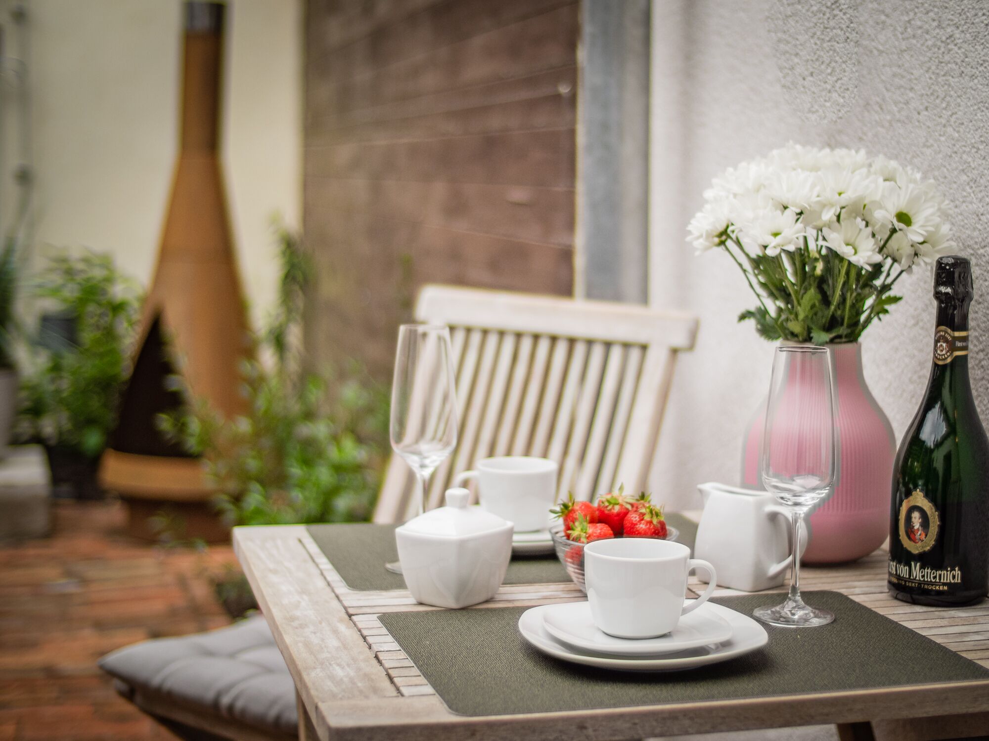 Une table dressée avec des tasses blanches, un verre, une bouteille et un vase contenant des fleurs blanches.