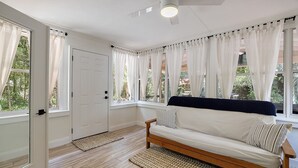 Una habitación con piso de madera, paredes blancas, una puerta blanca y un sofá blanco con una manta oscura, y vistas a zonas verdes a través de las ventanas.