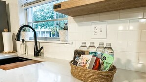 Una cocina con encimera blanca, un lavamanos, una ventana con plantas y una cesta con botellas de agua y una tarjeta.