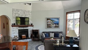 A living room with a stone fireplace, a sofa set, a dining table with chairs, and a television mounted above the fireplace. - Luxury Cottage in Alex Bay 3 bedrooms, 2.5 baths.  Book your 2026 Vacation Now!! (Alexandria Bay)