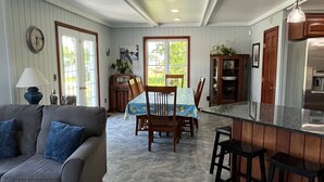 A spacious living room with a grey sofa, a dining area with a table and chairs, and a kitchen area with a countertop and bar stools. - Luxury Cottage in Alex Bay 3 bedrooms, 2.5 baths.  Book your 2026 Vacation Now!! (Alexandria Bay)