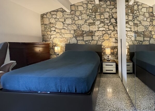 A bedroom with a stone wall, a bed with a blue bedspread, two bedside tables with lamps, and a wooden dresser.