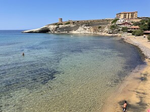 A coastal beach with clear water, a sandy shore, and a building on a hill.