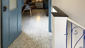 Un couloir étroit avec une porte bleue qui donne sur une pièce dont un des murs est en pierre et qui contient un lit.
