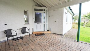 A covered patio with two black chairs, a white door, and a brick floor. - Limhus Cottage (Keswick)