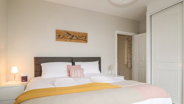 A neatly made bed with white linens and a yellow throw blanket, a wooden wall art piece above, and a closed white door to the right. - Limhus Cottage (Keswick)