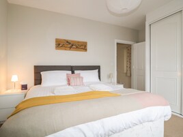 A neatly made bed with white linens and a yellow throw blanket, a wooden wall art piece above, and a closed white door to the right.