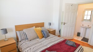 A neatly arranged bedroom with a bed, bedside tables, a lamp, a red throw blanket, and a view of the bathroom. - 1 Closheen Lane (Rosscarbery)