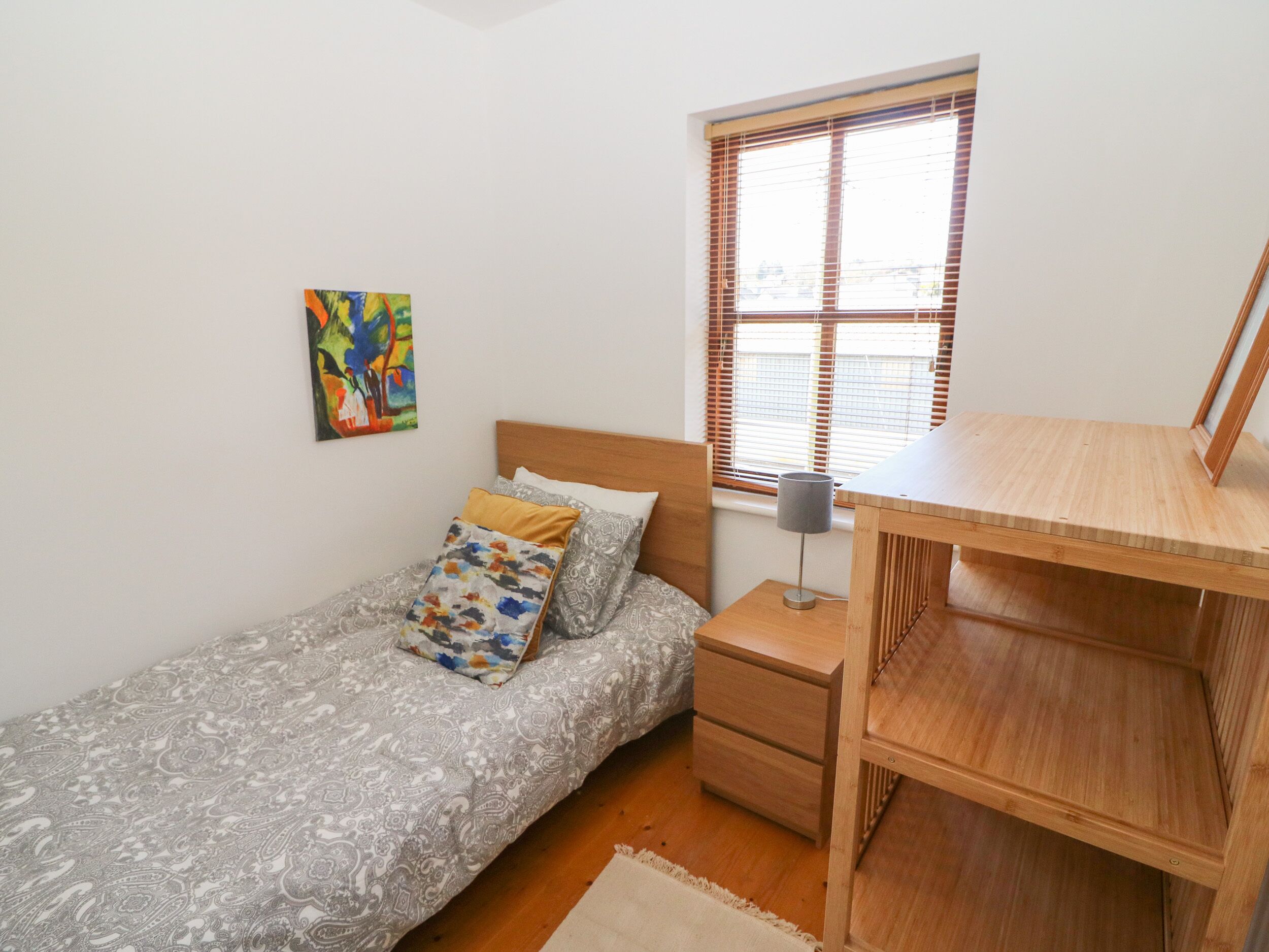 A small bedroom with a bed, a bedside table, a desk, a window with blinds, and a colorful painting on the wall.