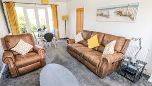 A living room with two brown leather armchairs, a grey oval table, and a brown sofa with yellow cushions. There is a dining table with chairs and a floor lamp. A painting is on the wall. - 53 St. Brides View (Haverfordwest)