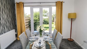 A dining area with a glass table set for four, grey upholstered chairs, a vase with white flowers, and a view of the garden through large windows. - 53 St. Brides View (Haverfordwest)