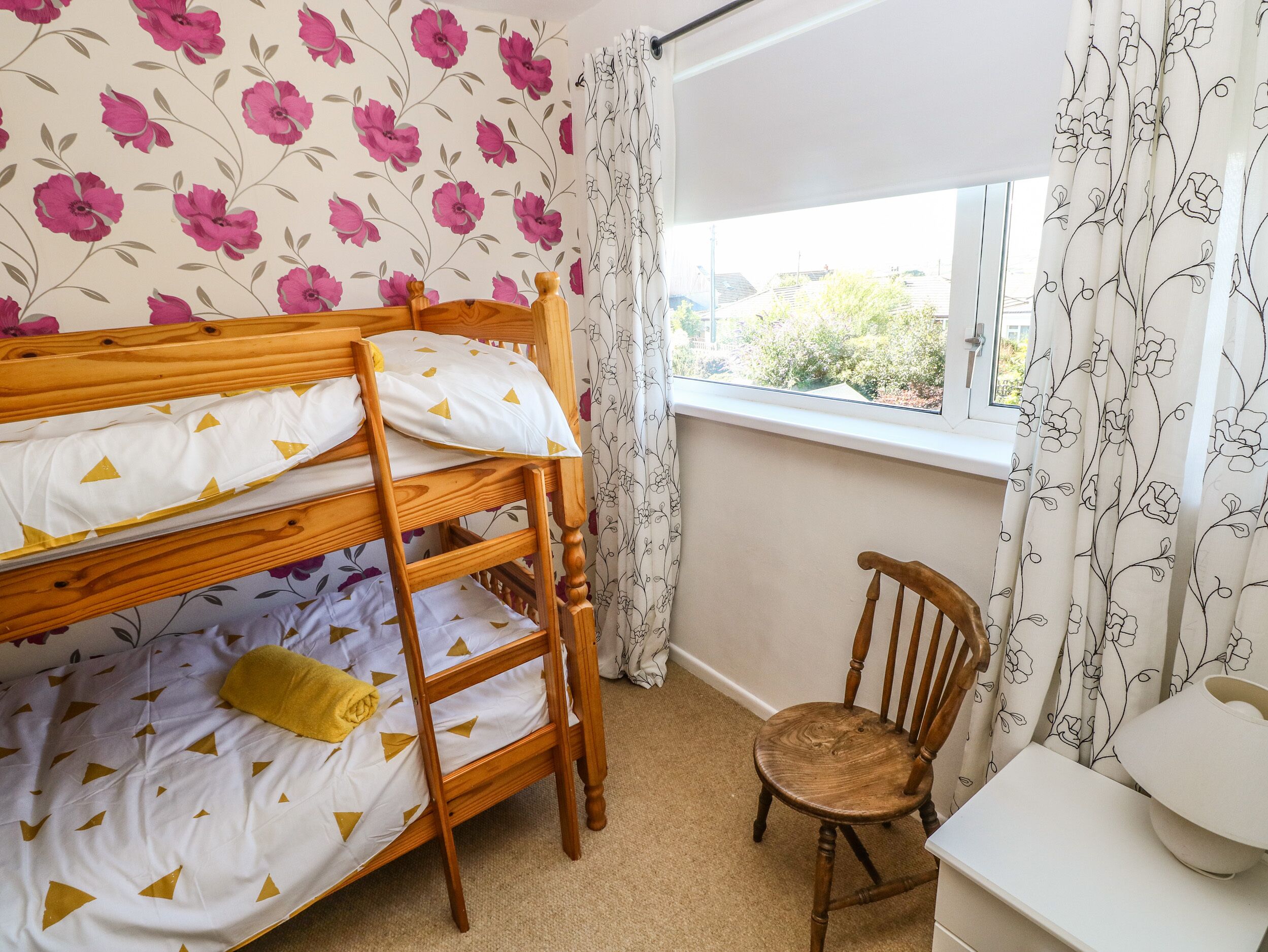 A bunk bed with a wooden frame, a chair, and a window with floral curtains.