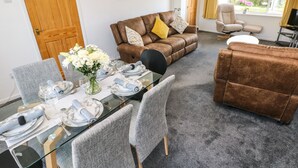 A furnished living room with a brown sofa, a glass dining table set for four, grey upholstered chairs, and a large window with curtains. - 53 St. Brides View (Haverfordwest)