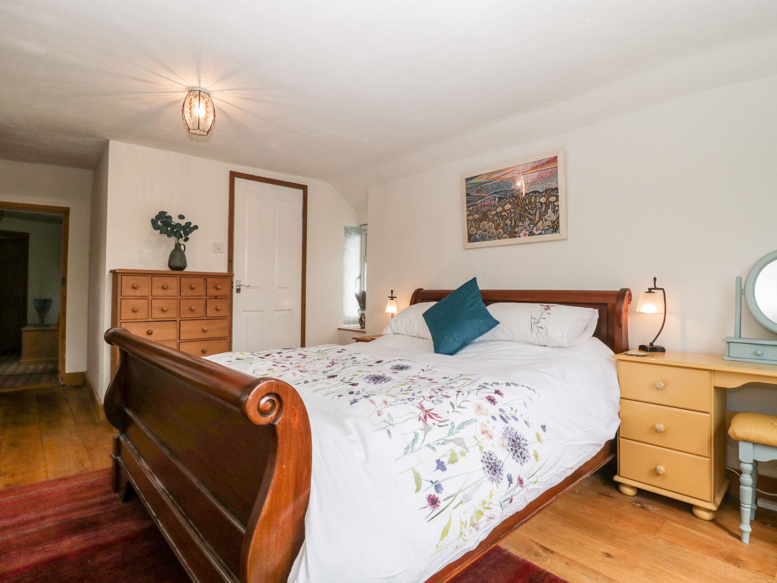 A bedroom with a wooden bed, a nightstand with a lamp, a dresser, and a painting on the wall.