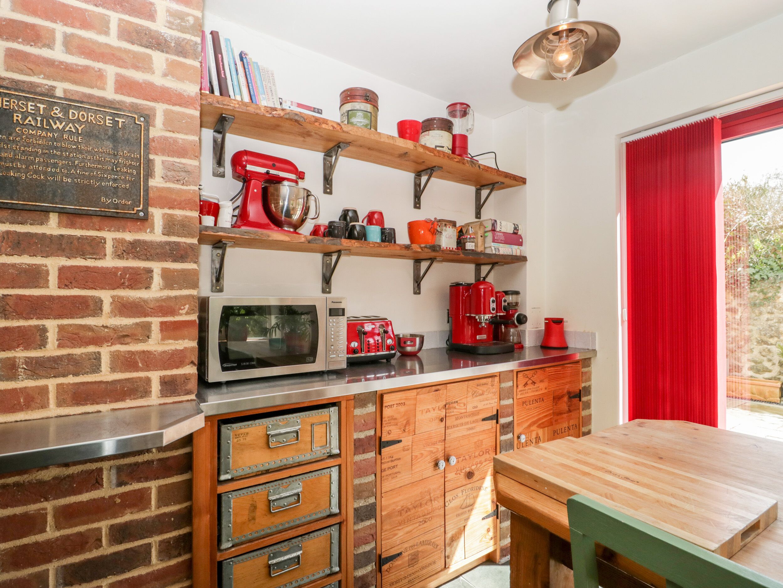 A kitchen with a brick wall, wooden shelves, a red mixer, a microwave, a coffee maker, and a wooden table.