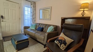 Ein Wohnzimmer mit einem braunen Ledersofa, einem passenden Sessel, einem kleinen blauen Truhen und einer Lampe auf einem Holztisch.