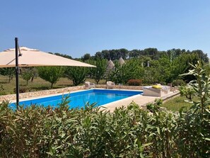 A swimming pool with a beige umbrella, surrounded by greenery and stone structures.