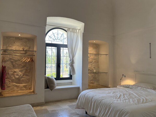 A bedroom with a large bed, a window with curtains, a wardrobe with hanging clothes, and a stone wall.