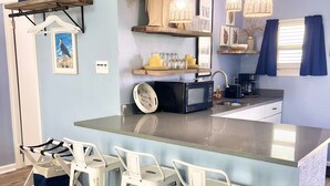 A modern kitchen with a central island, bar stools, a microwave, a coffee maker, and decorative items on shelves. - The Mark at Surfside, Apt. 9 (Surfside Beach)
