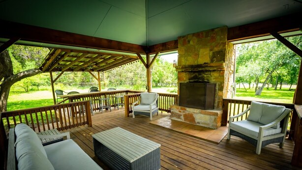 A covered outdoor patio with wooden flooring, white wicker furniture, and a stone fireplace.