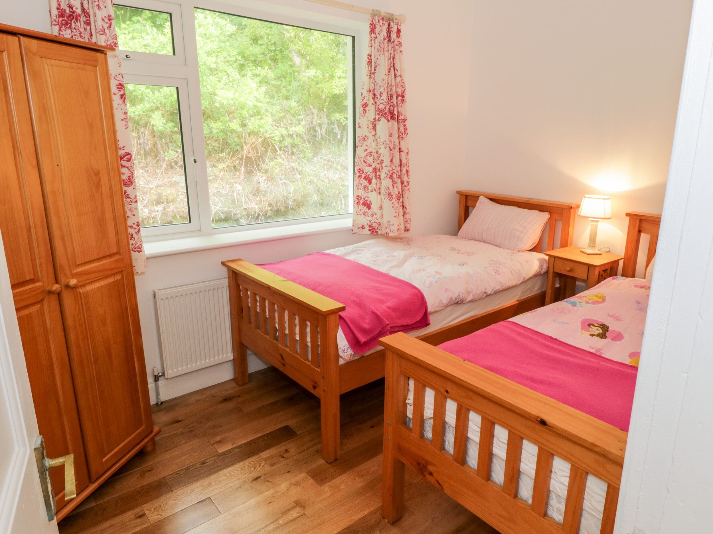 A bedroom with two wooden beds, a window with floral curtains, a wooden wardrobe, and a radiator.