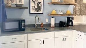 Une cuisine moderne avec des meubles blancs, un évier, un four à micro-ondes et une machine à café. Des objets décoratifs sont disposés sur les étagères et un tableau encadré orne le mur.