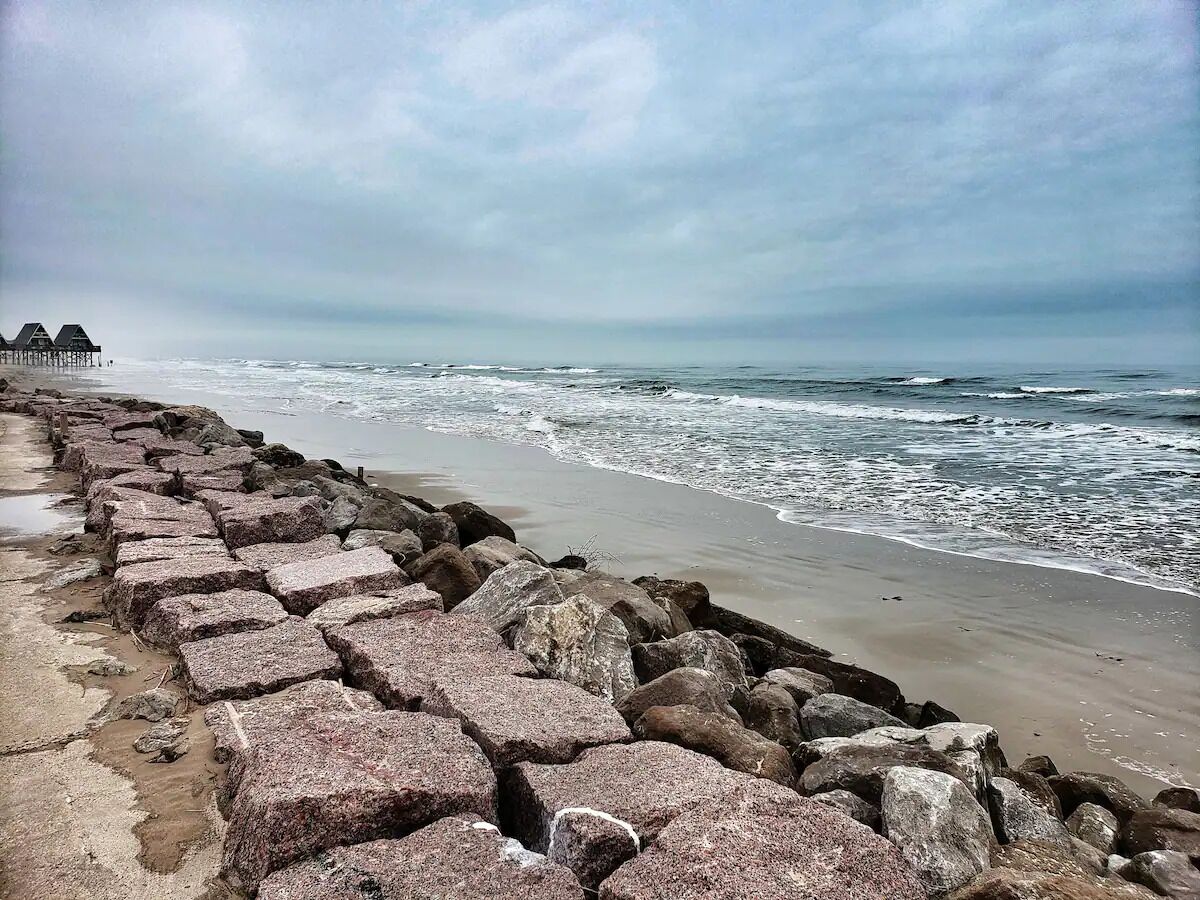 Una playa con un rompeolas rocoso y una hilera de chozas al fondo.