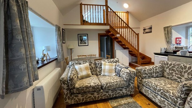 A living room with two patterned sofas, a wooden staircase, a dining table, and a kitchen area. - Talla Hope (Kilmuir)