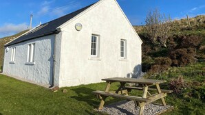 A small, single-story building with a white exterior and a dark roof, situated on a grassy area with a picnic table nearby. - Talla Hope (Kilmuir)