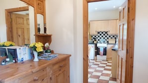 A hallway leading to a kitchen with a checkered floor, wooden cabinets, and a mirror above a wooden dresser with books and flowers. - Borahard Lodge (Naas)