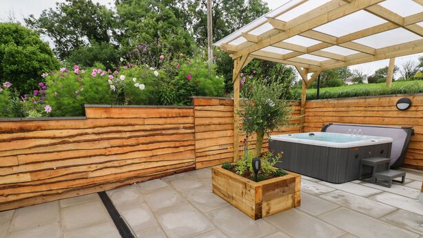 A patio with a wooden pergola, a hot tub, and a planter box with a tree. - Brynawel (Carmarthen)