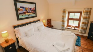 A neatly made bed with white linens, a wooden headboard, and a painting of a coastal town above it. There are two bedside lamps, a window with curtains, and a small wooden nightstand with drawers. - Brynawel (Carmarthen)