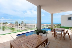 Un espacio al aire libre techado con piscina, mobiliario de madera y vista al mar.