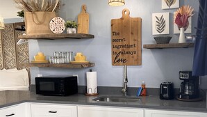 Eine moderne Küche mit weißen Schränken, einer schwarzen Mikrowelle, einem Spülbecken und einer Kaffeemaschine. An der Wand hängt ein dekoratives Schild und auf dem Regal steht ein Korb.