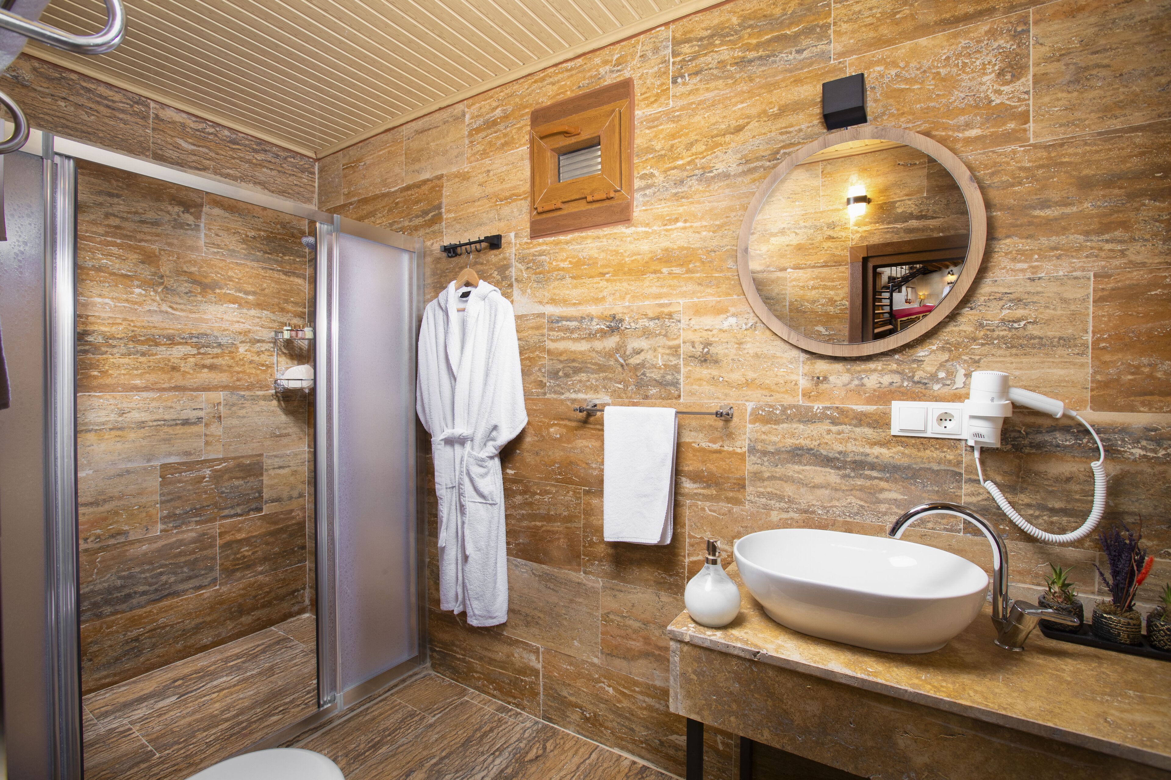 Un baño moderno con pared de piedra, un espejo redondo, un lavabo blanco y una ducha con cerramiento de vidrio.