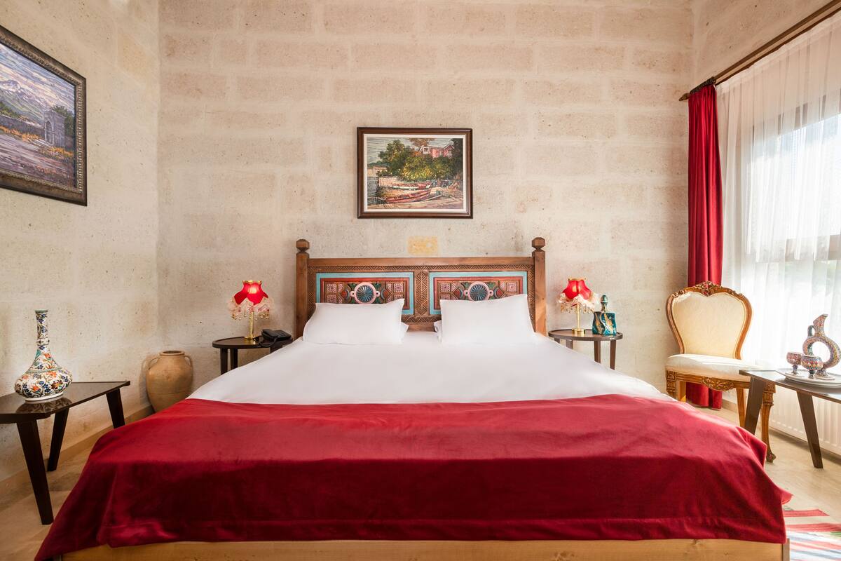 Habitación de hotel con una cama grande, dos mesitas de noche, una silla, una cortina roja y un cuadro en la pared.