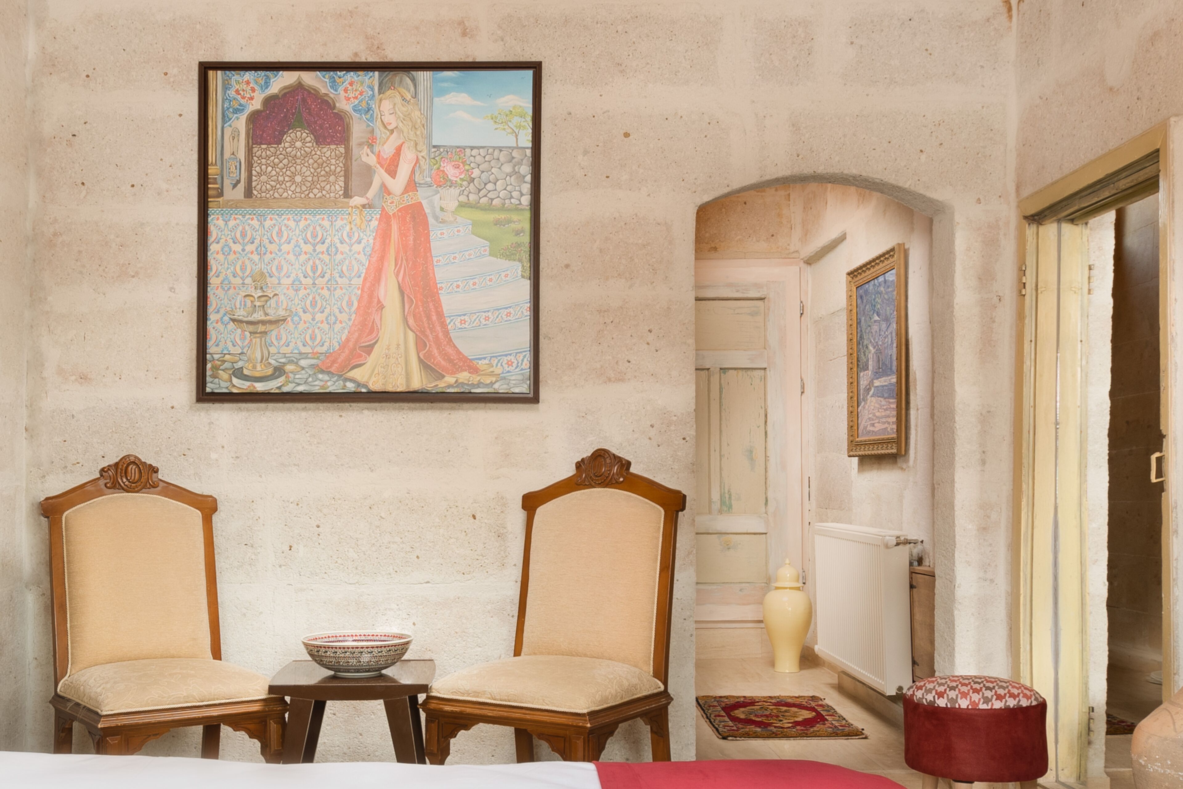 Una habitación con dos sillas de madera, una mesa con un cuenco, un cuadro en la pared y una puerta que da a otra habitación.