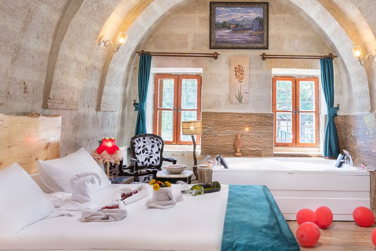 Habitación de hotel con una cama grande, un rincón de estar con una silla, una mesita y vistas al exterior a través de dos ventanas.