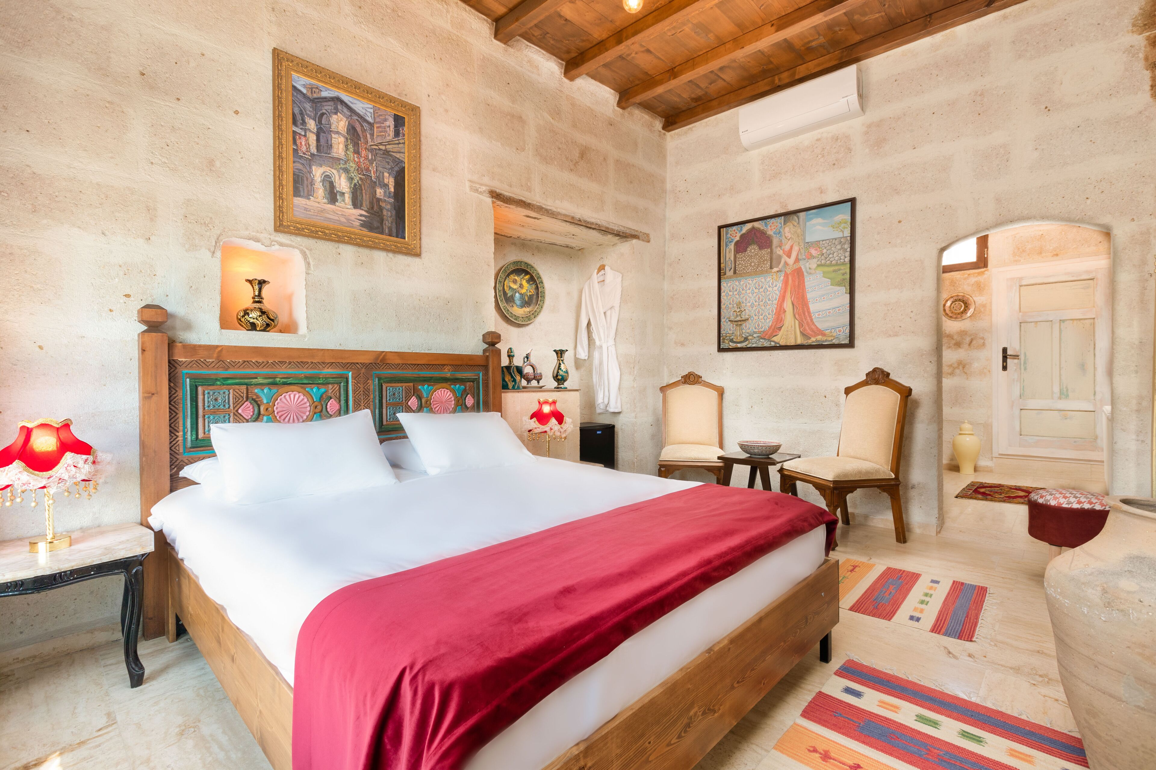 A bedroom with a wooden bed, a red blanket, a nightstand with a lamp, a chair, a small table, and a door.
